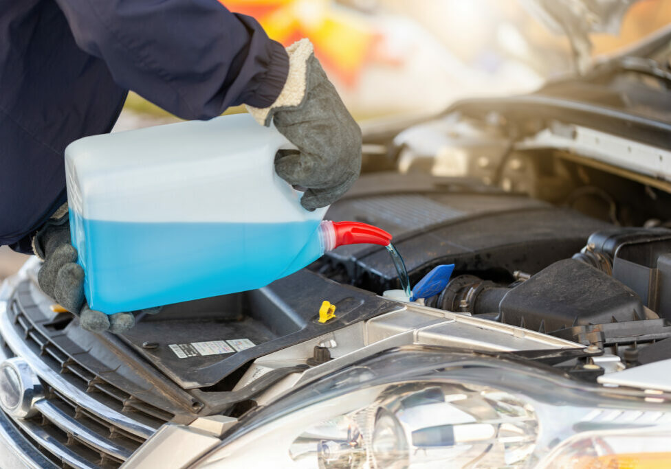 Person pouring antifreeze to the car in wintertime. Technician with open hood adding cooland to the motor in winter. Human hand with bottle of blue liquid.